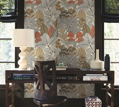 A wooden desk with books, a lamp, and vases sits in front of York Wallcoverings’ Namiri Wallpaper. A wooden chair is at the desk, and two large windows with black curtains are in the background.