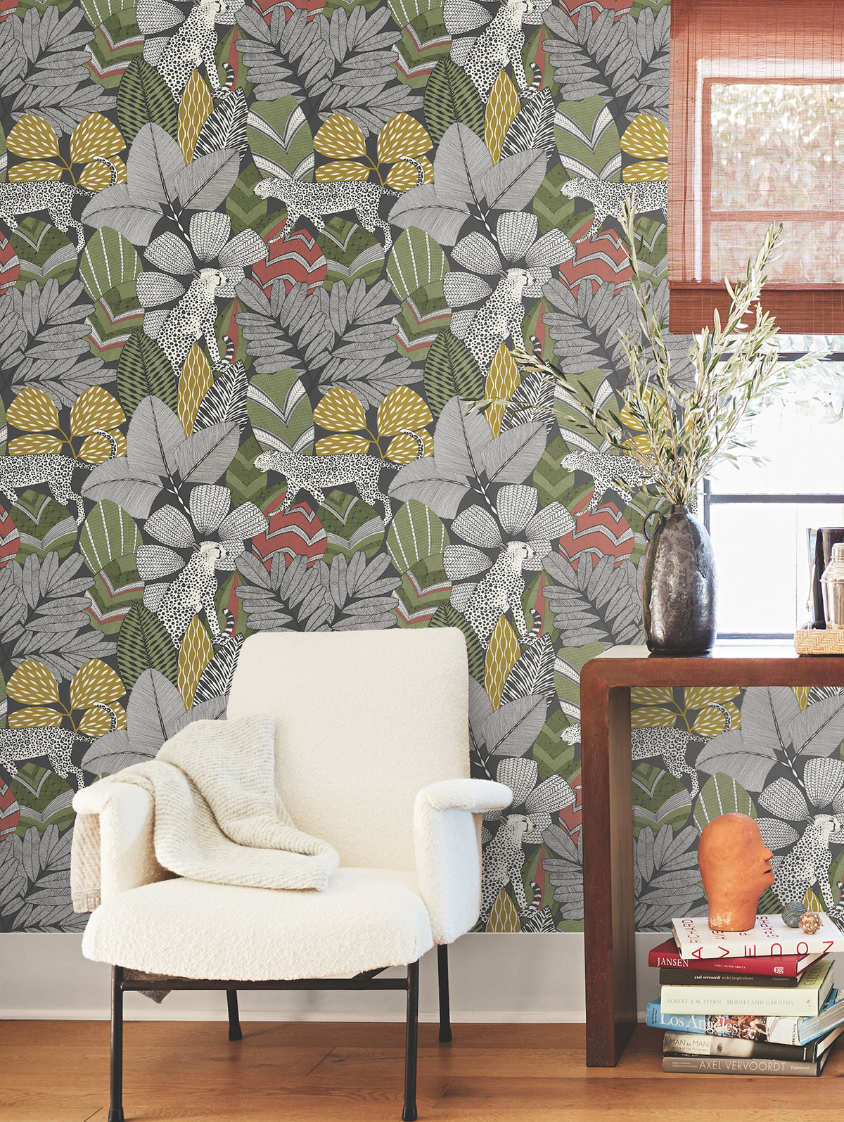 A cozy white armchair with a blanket sits beside a wooden table holding a vase with greenery, books, and glasses, set against York Wallcoverings’ Namiri Wallpaper and a window with a bamboo shade.