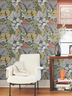 A cozy white chair with a blanket sits by a wooden table holding a vase with branches and stacked books, in front of the Namiri Wallpaper from York Wallcoverings, featuring its colorful floral pattern. A window with a bamboo shade is in the background.
