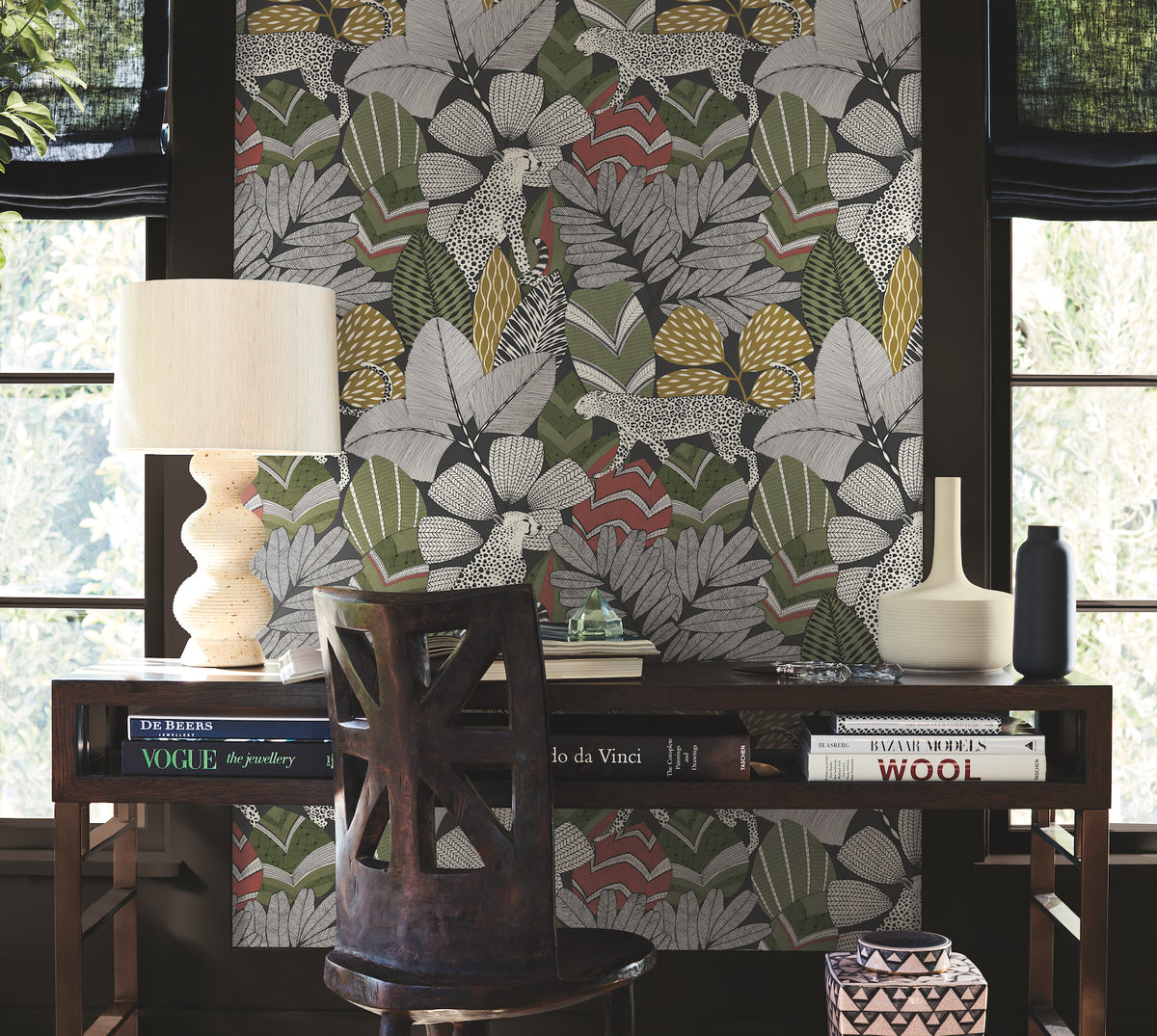 A wooden desk adorned with books, a lamp, and decorative accents sits in front of a wall covered in Namiri Wallpaper by York Wallcoverings, which features bold, colorful leaves and leopards. Two large windows with black shades let in natural light.