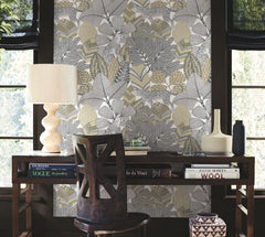 A desk adorned with art books, a sculpted lamp, vases, and a chair is set against a wall covered in York Wallcoverings’ Namiri Wallpaper—a bold, modern leaf-patterned design—while large windows fill the space with natural light.