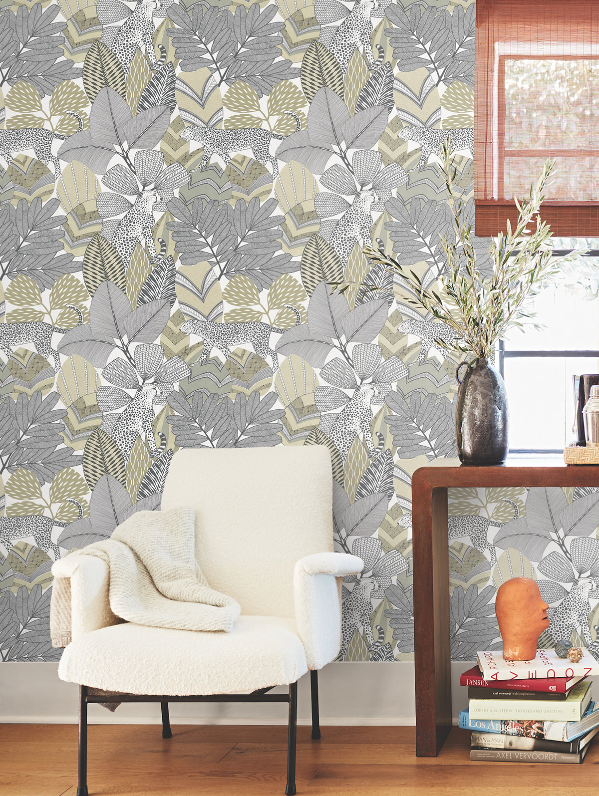 A cozy white armchair with a blanket sits by a wooden table, which holds a vase of leafy branches and stacked books, set against a wall adorned with York Wallcoverings’ Namiri Wallpaper in a grey and yellow leafy pattern. Sunlight streams through a window dressed with a brown shade.