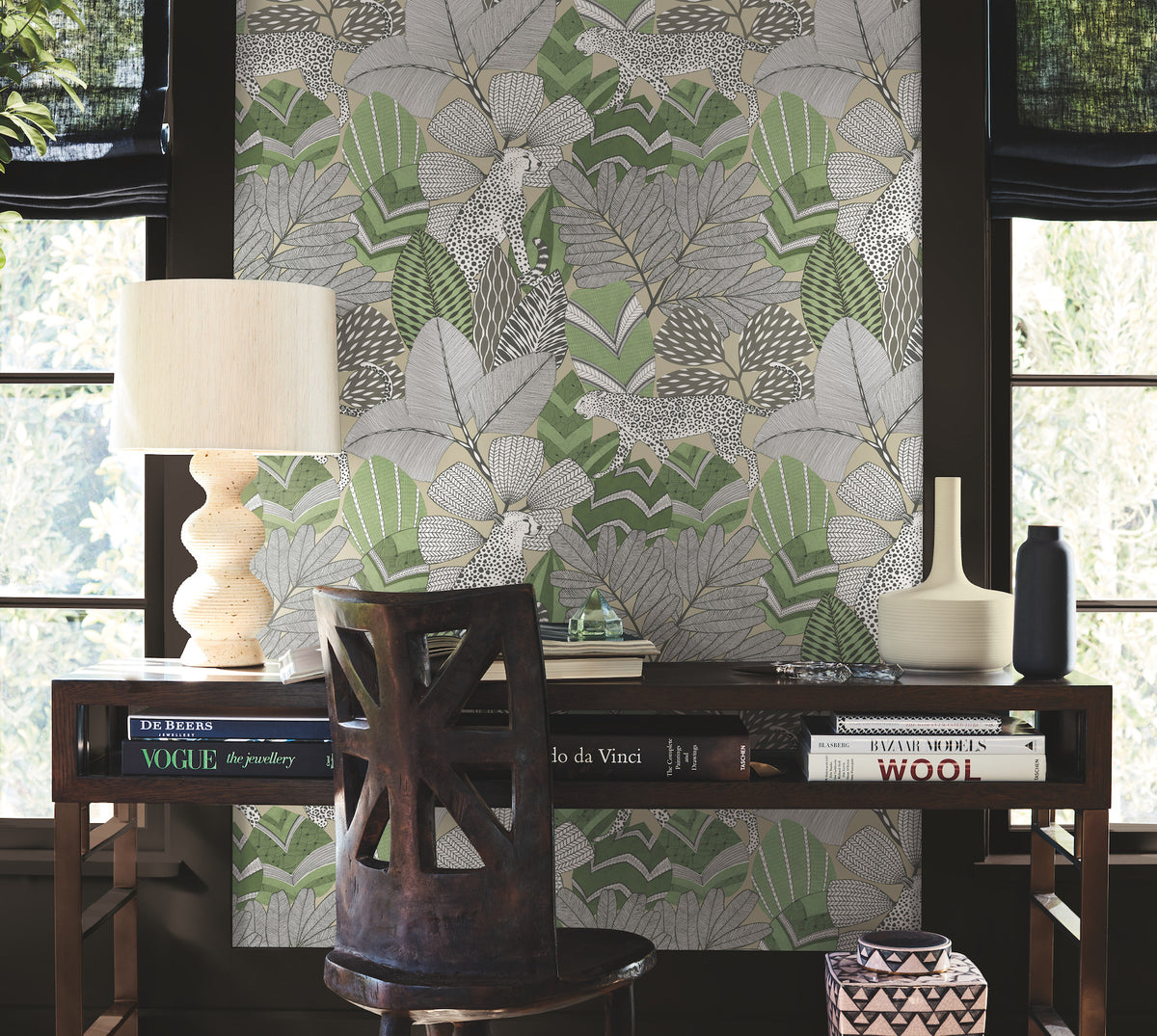 A wooden desk with books, a lamp, and ceramics sits in front of a window, accompanied by a chair. The wall behind is adorned with York Wallcoverings’ Namiri Wallpaper, featuring bold green and gray leaf patterns.