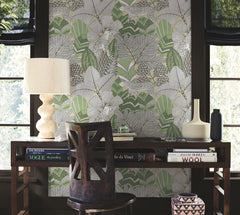 A wooden desk with books, a lamp, and ceramics sits in front of a window, accompanied by a chair. The wall behind is adorned with York Wallcoverings’ Namiri Wallpaper, featuring bold green and gray leaf patterns.