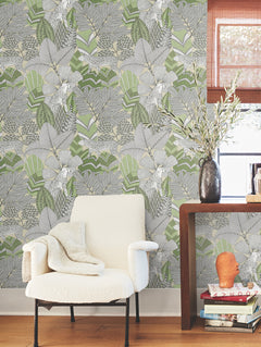 A cozy white chair with a blanket sits beside a wooden table holding books, a vase with branches, and a window; the wall features the Namiri Wallpaper by York Wallcoverings, showcasing its leafy green and gray botanical pattern.