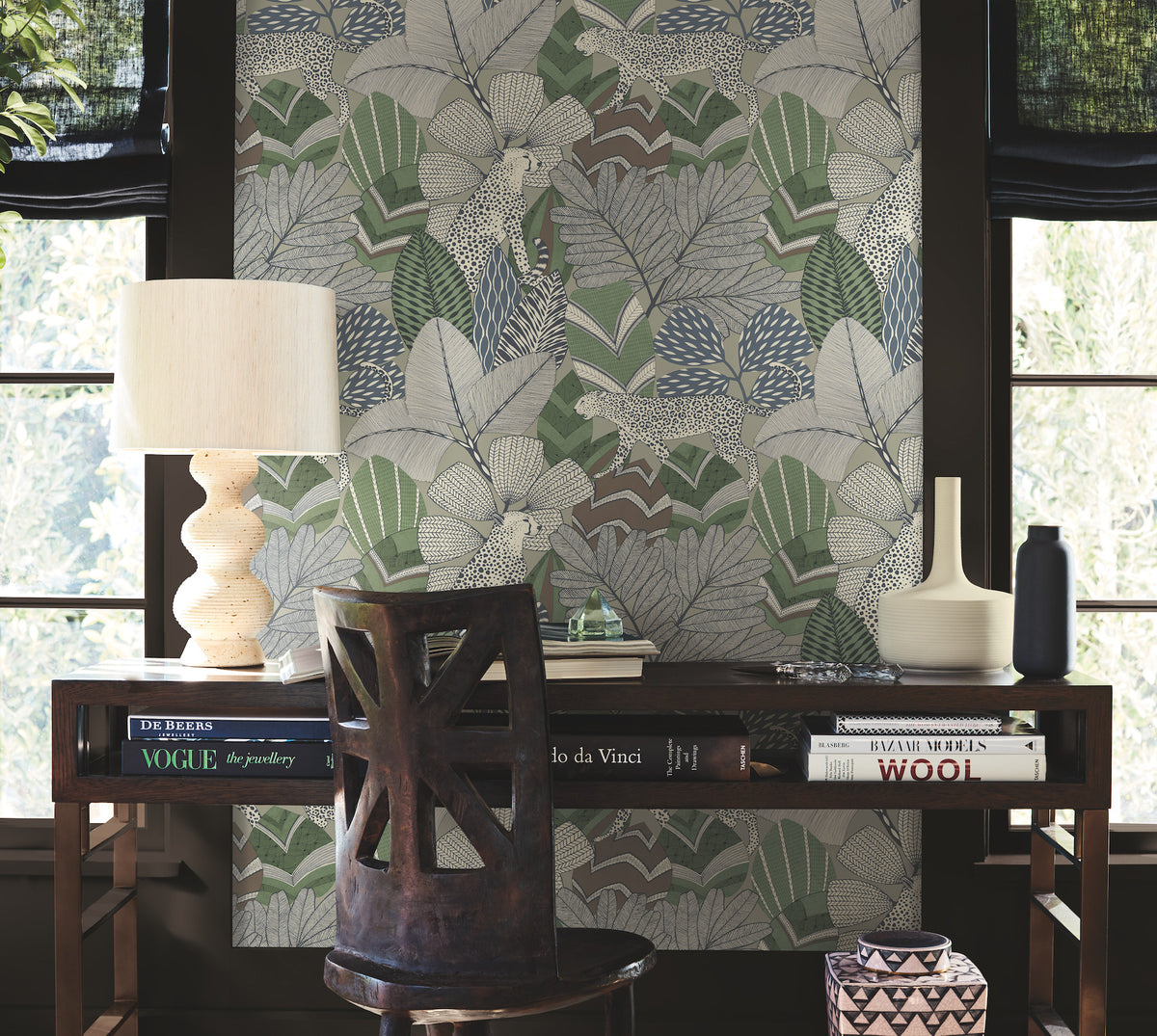 A wooden desk adorned with books, a lamp, and vases sits in front of a window featuring Namiri Wallpaper by York Wallcoverings with its leafy design. A wooden chair is placed at the desk as sunlight filters in from outside.