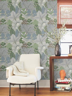 A cozy white armchair with a blanket sits near a small wooden table holding books and a vase with greenery, set against York Wallcoverings’ Namiri Wallpaper, which features a striking large leafy green and gray botanical pattern.