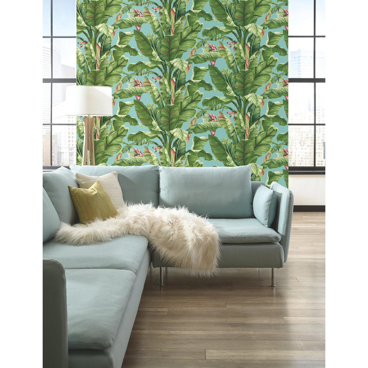 Modern living room with light blue sectional sofa, white fur throw, and green pillow. The wall features Banana Leaf Wallpaper by York Wallcoverings with a green tropical leaf pattern. Large windows reveal a cityscape backdrop, and a white floor lamp stands nearby.
