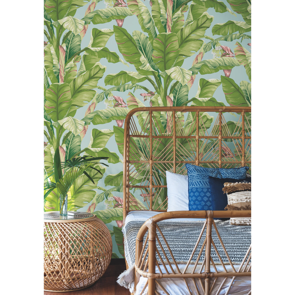 A rattan bed and side table stand in front of the Banana Leaf Wallpaper by York Wallcoverings. The bed is adorned with patterned blue and gray pillows and a woven blanket, creating a cozy, natural, and tropical-themed bedroom scene.