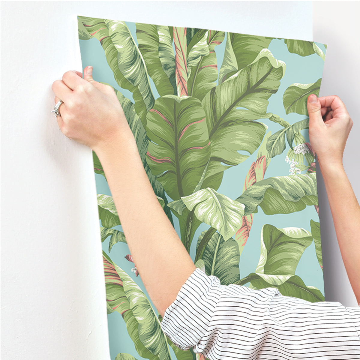 A person in a striped shirt applies York Wallcoverings’ Banana Leaf Wallpaper to a white wall, with only their hands and forearms visible.