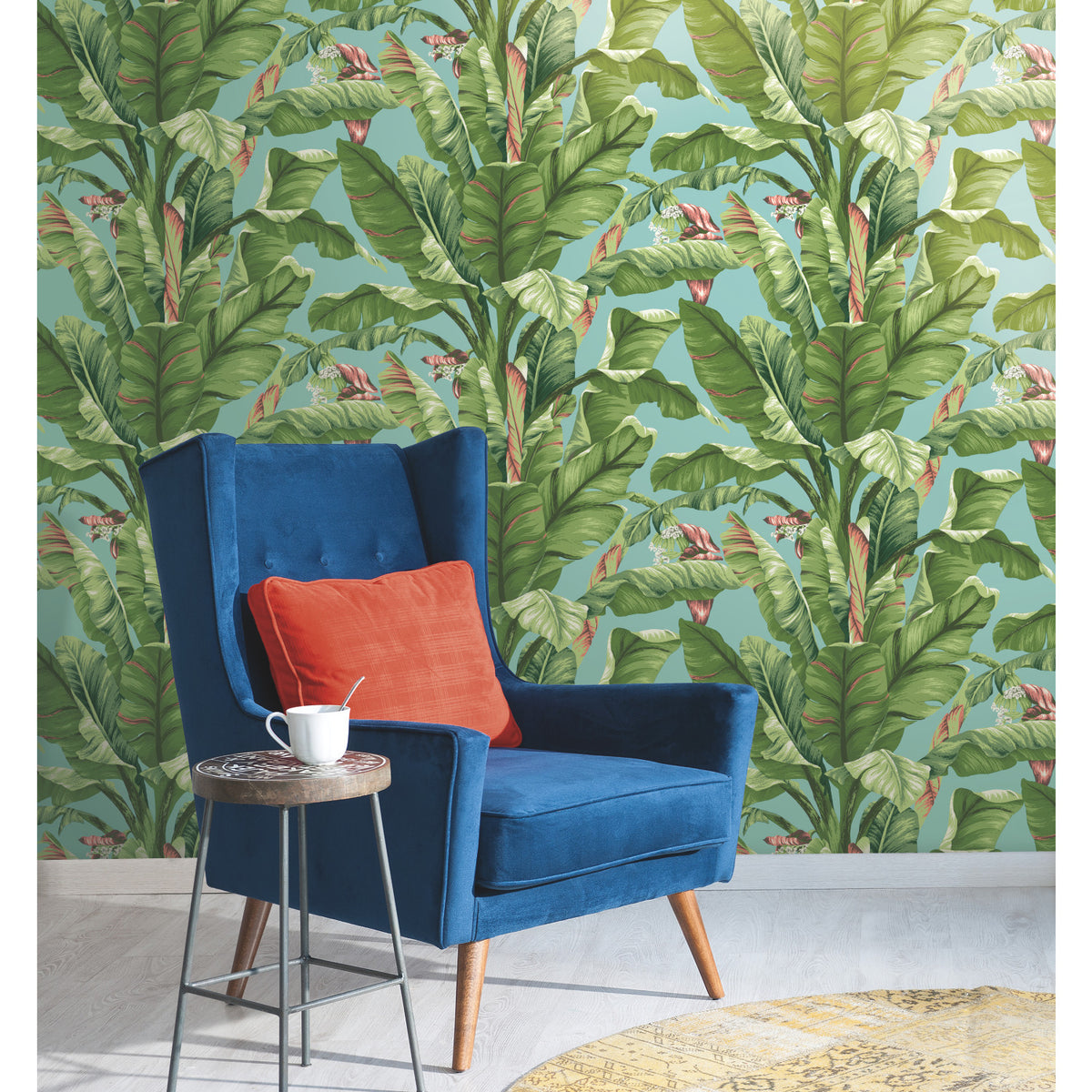 A blue armchair with an orange cushion sits beside a small table holding a white mug, set against York Wallcoverings’ Banana Leaf Wallpaper and a round yellow rug on a light wood floor.