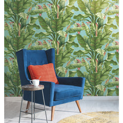 A blue armchair with an orange cushion sits beside a small table holding a white mug, set against York Wallcoverings’ Banana Leaf Wallpaper and a round yellow rug on a light wood floor.