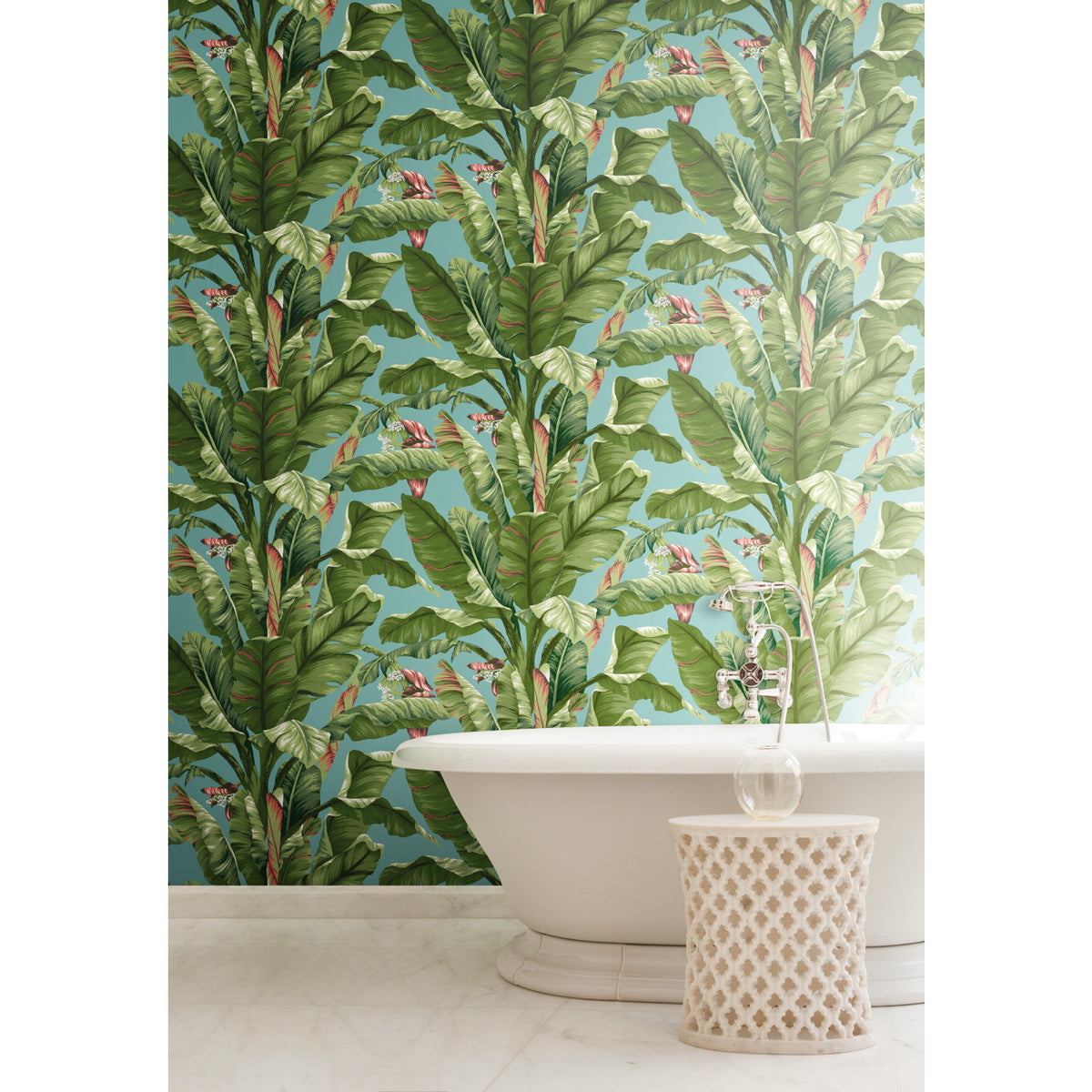 A white bathtub and a small round table are positioned in front of the York Wallcoverings Banana Leaf Wallpaper, which features green tropical leaves and pink flowers on a light blue background. The floor is finished in white marble.