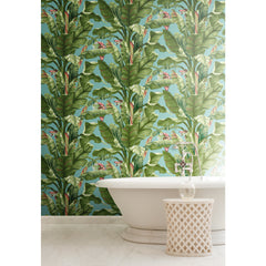 A white bathtub and a small round table are positioned in front of the York Wallcoverings Banana Leaf Wallpaper, which features green tropical leaves and pink flowers on a light blue background. The floor is finished in white marble.