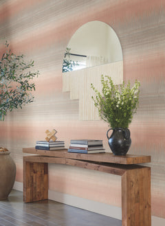 A wooden console table with stacked books, a black vase of white flowers, and decor sits against a wall covered in York Wallcoverings’ Desert Textile Wallpaper, featuring pink and gray stripes. Above the table is a large, uniquely shaped mirror, and a leafy plant stands on the left.