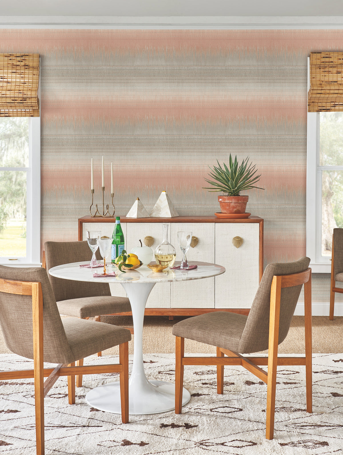 A mid-century modern dining area featuring a round white table, four brown chairs, and a sideboard adorned with decor and a potted plant. The walls are covered in York Wallcoverings’ Desert Textile Wallpaper, while two windows are outfitted with bamboo shades.