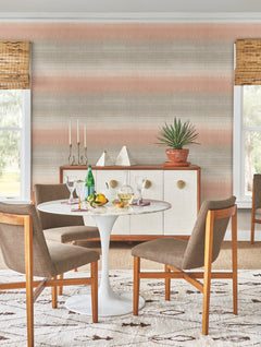 A mid-century modern dining area featuring a round white table, four brown chairs, and a sideboard adorned with decor and a potted plant. The walls are covered in York Wallcoverings’ Desert Textile Wallpaper, while two windows are outfitted with bamboo shades.