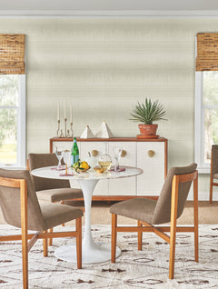 A modern dining room features a round white table with four brown chairs, drinks, lemons, and plants. Along the desert textile wallpaper by York Wallcoverings sits a sideboard with decor, positioned between two large windows with bamboo shades.