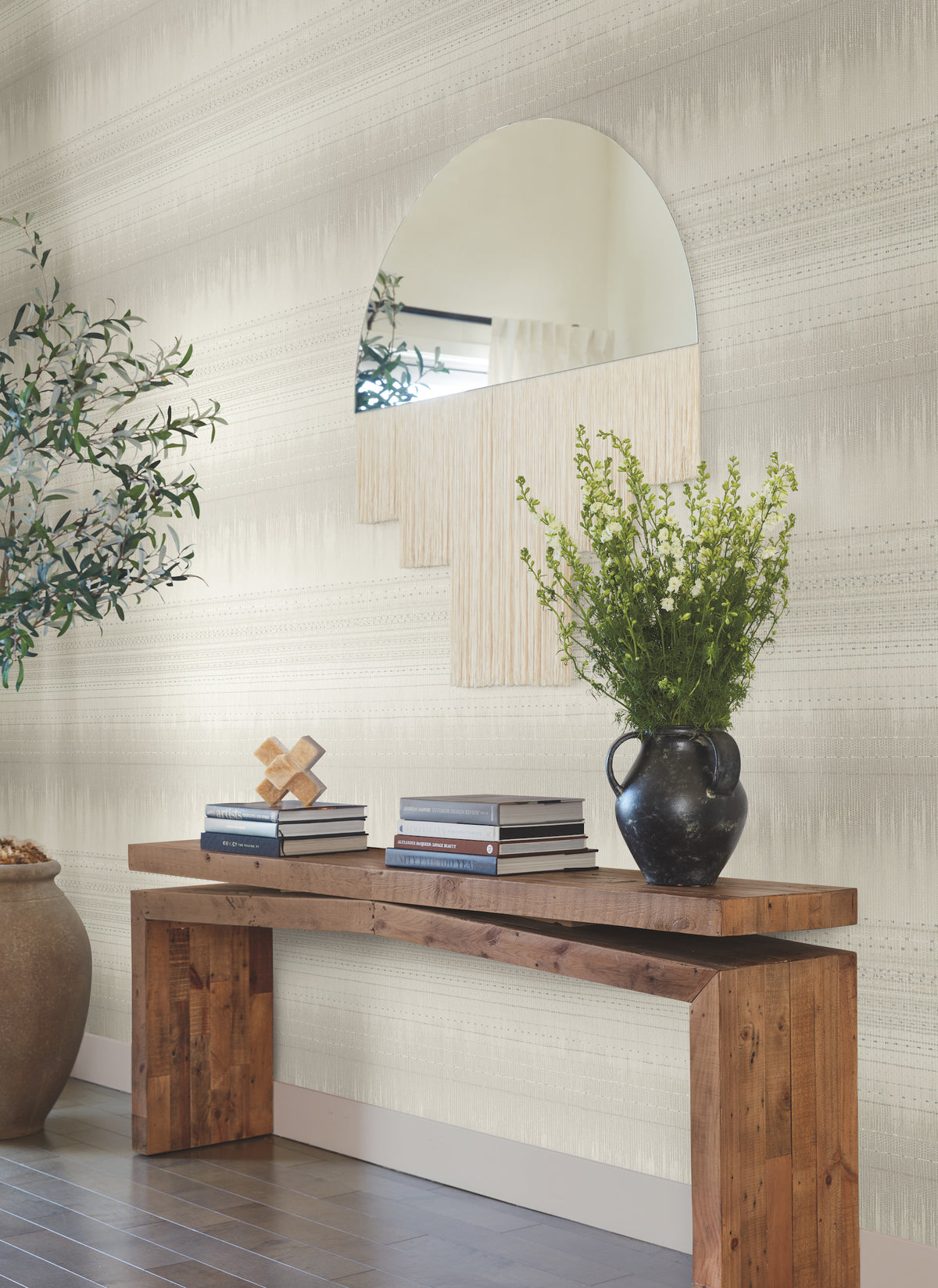 A wooden console table with stacked books, a black vase filled with green foliage, and a decorative sculpture sits below a large, uniquely-shaped mirror on a wall adorned with York Wallcoverings’ Desert Textile Wallpaper. An indoor tree stands on the left side.