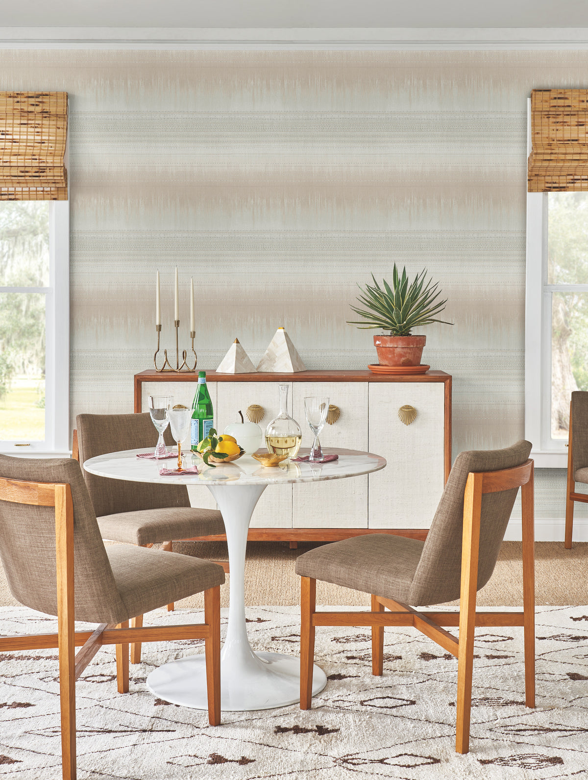 A mid-century modern dining area featuring a round white table, four brown chairs, drinks, fruit, and a sideboard topped with decor and a potted plant is set against York Wallcoverings’ Desert Textile Wallpaper. Two windows with bamboo shades frame the scene.