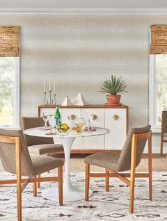 A mid-century modern dining area featuring a round white table, four brown chairs, drinks, fruit, and a sideboard topped with decor and a potted plant is set against York Wallcoverings’ Desert Textile Wallpaper. Two windows with bamboo shades frame the scene.