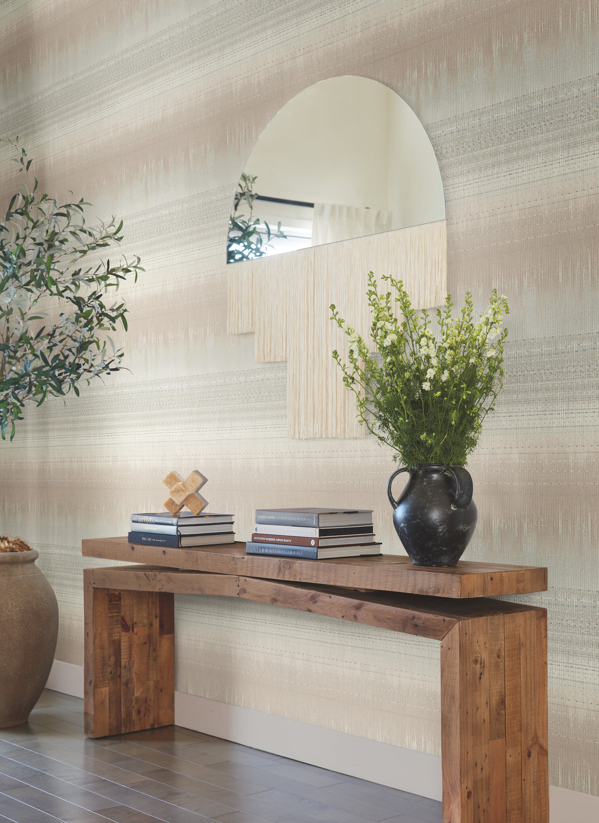 A rustic wooden console table with books, a black vase of green flowers, and decorative objects sits against a wall adorned with York Wallcoverings’ Desert Textile Wallpaper. Above the table, a geometric mirror reflects part of the room, while a large potted plant stands nearby.