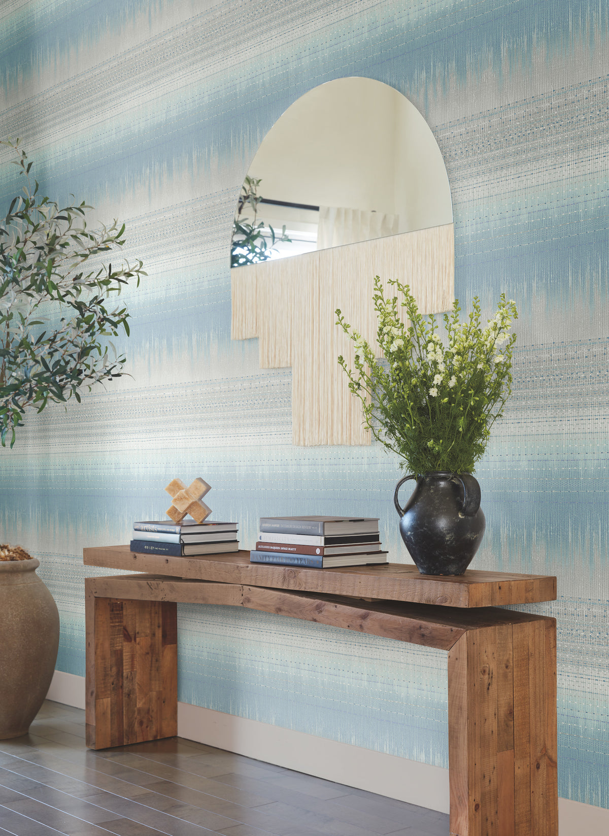 A wooden console table with books and a black vase of flowers sits against a wall adorned with York Wallcoverings’ Desert Textile Wallpaper. Above the table is a large, uniquely shaped mirror, and an olive tree in a pot is visible on the left.