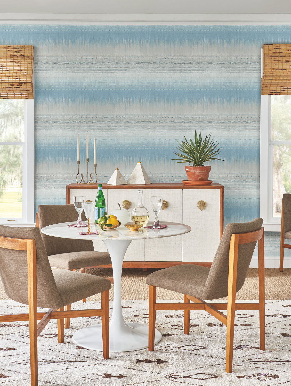 A round white table with drinks, fruit, and glasses sits with four wooden chairs. Behind, there’s a sideboard adorned with plants, decor, and a candelabra against York Wallcoverings’ Desert Textile Wallpaper—a blue patterned backdrop—framed by two windows dressed in bamboo blinds.