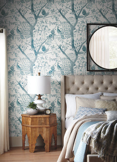 A cozy bedroom featuring York Wallcoverings’ Peacock Block Print Wallpaper in a soothing blue tree pattern, a tufted bed adorned with textured pillows, and a wooden nightstand displaying a lamp and plant. A round mirror hangs above the bed while sunlight streams through sheer curtains.