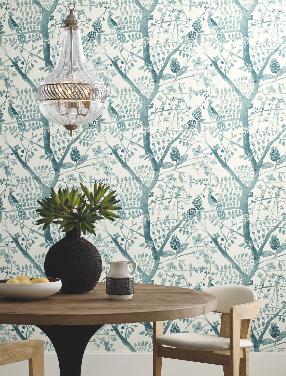 Round wooden table with a black vase of greenery, ceramic pitcher, and bowl. Cream upholstered chair, glass chandelier, and Peacock Block Print Wallpaper by York Wallcoverings featuring blue birds and trees in the background.