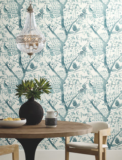 Round wooden table with a black vase of greenery, ceramic pitcher, and bowl. Cream upholstered chair, glass chandelier, and Peacock Block Print Wallpaper by York Wallcoverings featuring blue birds and trees in the background.