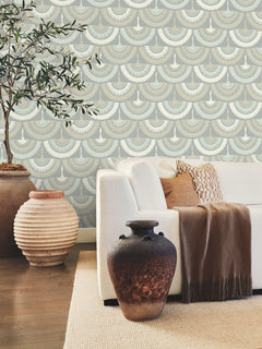 A white sofa with tan and patterned pillows, a brown throw blanket, and two large pottery vases sits in front of a wall adorned with York Wallcoverings’ Feather & Fringe Wallpaper. Behind the sofa, a leafy plant stands on the wooden floor.
