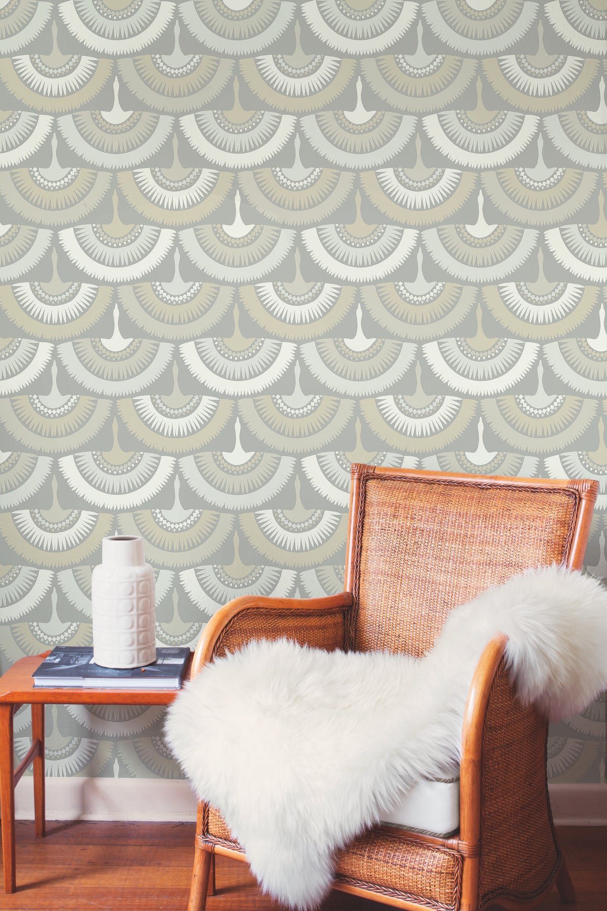 A wicker chair draped with a white faux fur throw sits beside a small side table topped with a white vase and two books. The background showcases York Wallcoverings’ Feather & Fringe Wallpaper, featuring art deco-inspired scalloped designs in beige, gray, and white.