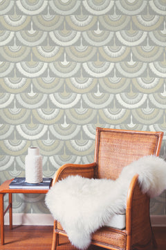 A wicker chair draped with a white faux fur throw sits beside a small side table topped with a white vase and two books. The background showcases York Wallcoverings’ Feather & Fringe Wallpaper, featuring art deco-inspired scalloped designs in beige, gray, and white.
