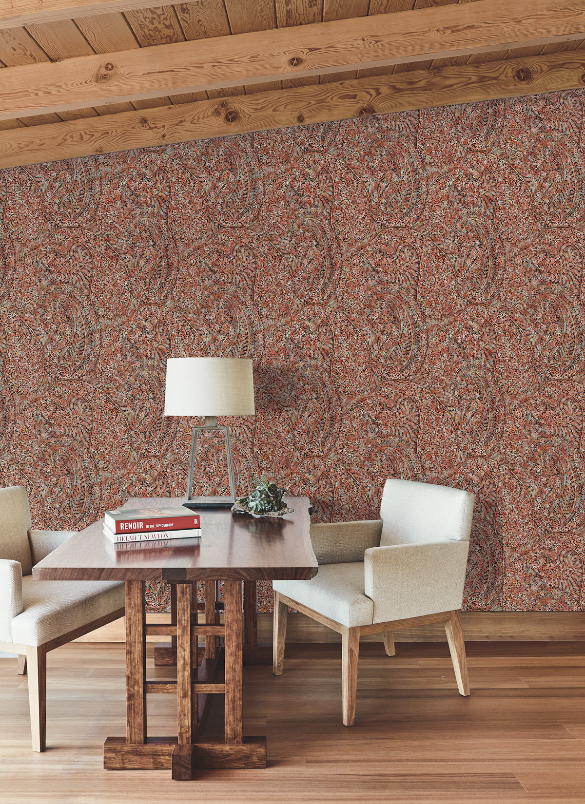 A wooden table topped with books, a lamp, and a plant sits between two white chairs in a room featuring wooden floors, wood beam ceilings, and Kashmir Dreams Paisley Wallpaper by York Wallcoverings in warm tones.