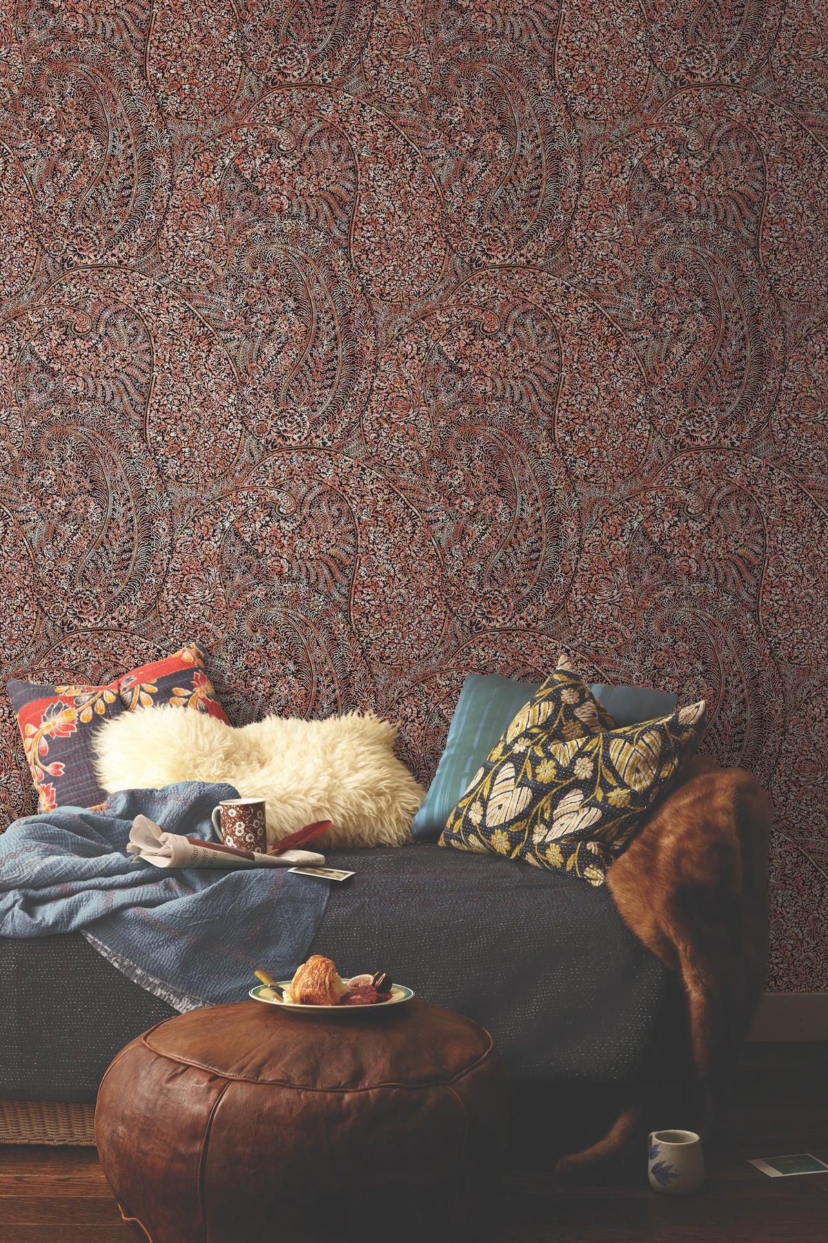 Cozy living space with a dark couch, assorted patterned pillows, a fur throw, a blanket, a mug, books, and a leather ottoman topped with a plate of pastries, set against York Wallcoverings’ Kashmir Dreams Paisley Wallpaper.