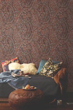 Cozy living space with a dark couch, assorted patterned pillows, a fur throw, a blanket, a mug, books, and a leather ottoman topped with a plate of pastries, set against York Wallcoverings’ Kashmir Dreams Paisley Wallpaper.