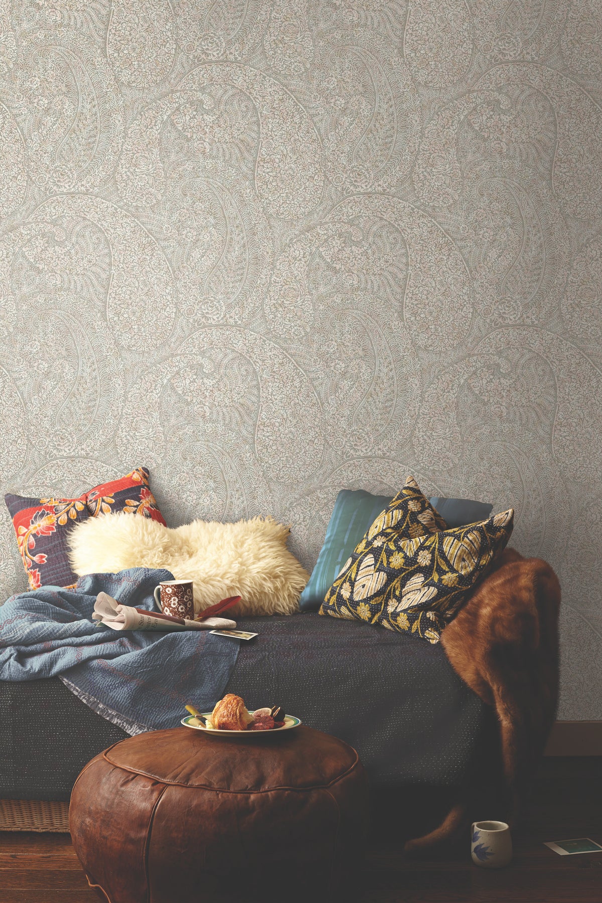 A cozy lounge area features patterned pillows, a fur throw, and a blue blanket draped over the sofa, beside a round leather ottoman topped with pastries and a mug. A cup and book rest on the wooden floor. The space is set against Kashmir Dreams Paisley Wallpaper by York Wallcoverings in the background.
