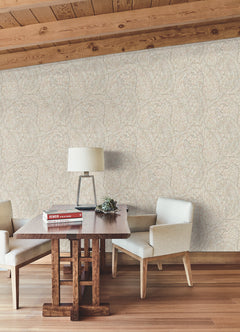 A wooden table with books, a lamp, and a plant sits between two cream chairs on a wood floor under a slanted wood ceiling, against a wallpapered wall featuring York Wallcoverings’ Kashmir Dreams Paisley Wallpaper.