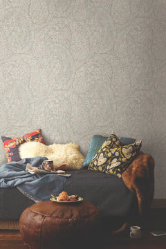 In a warmly lit, textured room adorned with York Wallcoverings’ Kashmir Dreams Paisley Wallpaper, a cozy sofa with patterned pillows, a sheepskin throw, mugs, a blue blanket, and a leather ottoman displaying pastries and a cup create an inviting scene.