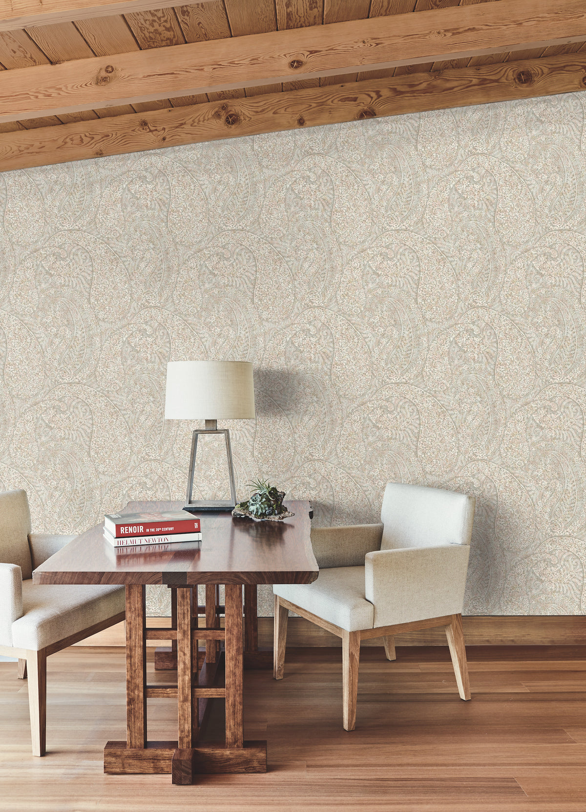 Sure! Here’s a revised sentence incorporating the provided product data: A small wooden table with books, a lamp, and a plant sits between two cream-colored chairs in a cozy room with wooden floors, a sloped wood-paneled ceiling, and Kashmir Dreams Paisley Wallpaper by York Wallcoverings.