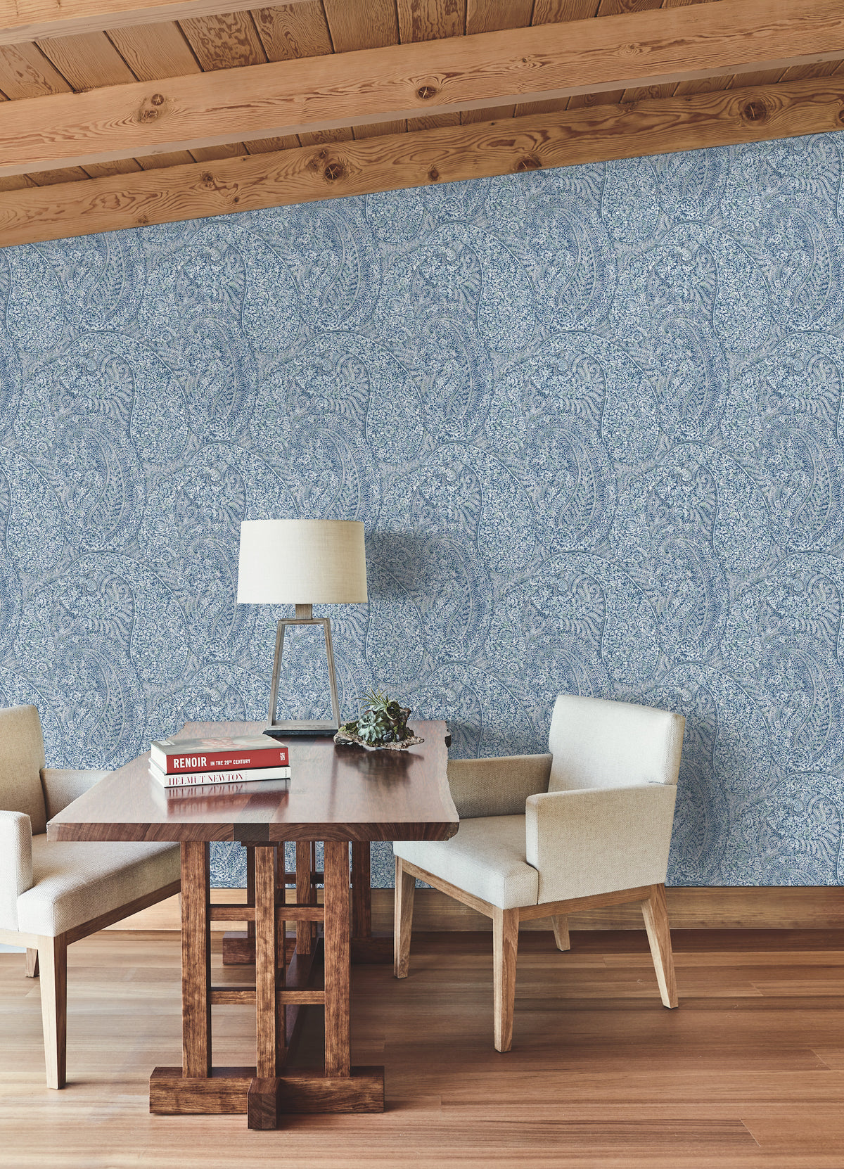 A small wooden table topped with books, a lamp, and a plant sits between two cream chairs in a room featuring Kashmir Dreams Paisley Wallpaper by York Wallcoverings, wooden floors, and a slanted wooden ceiling.