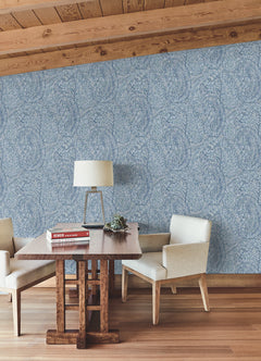 A wooden table adorned with a lamp, books, and a small plant is flanked by two cream chairs, set against York Wallcoverings’ Kashmir Dreams Paisley Wallpaper in blue, with a wooden floor beneath and a slanted wood ceiling overhead.
