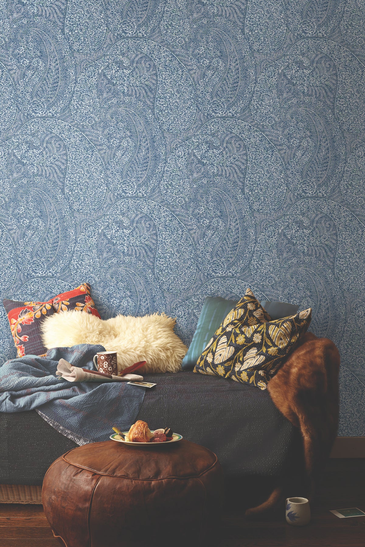 A cozy sofa with patterned pillows, a fur throw, a blanket, and mugs sits against the ornate Kashmir Dreams Paisley Wallpaper by York Wallcoverings. A leather ottoman holding a plate of snacks and a cup is placed in front, creating a warm and inviting scene.