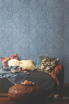 A cozy sofa with patterned pillows, a fur throw, a blanket, and mugs sits against the ornate Kashmir Dreams Paisley Wallpaper by York Wallcoverings. A leather ottoman holding a plate of snacks and a cup is placed in front, creating a warm and inviting scene.