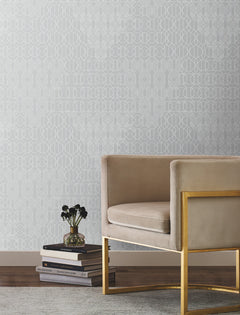 A stylish beige velvet chair with a gold frame is placed beside stacked books and a small black vase of flowers, set against the Modern Chandelier Wallpaper by York Wallcoverings, in a bright room featuring a light rug and wooden floor.