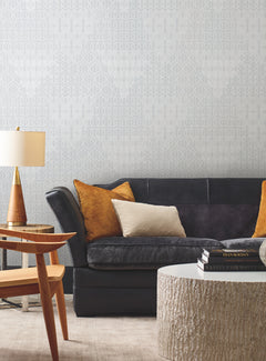 A modern living room featuring a dark gray sofa with yellow and beige pillows, a round textured coffee table, wooden side table, and lamp, complemented by light patterned Modern Chandelier Wallpaper from York Wallcoverings. Warm and neutral tones create a cozy, stylish atmosphere.