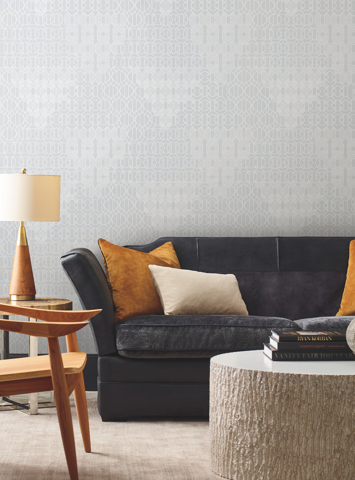 A modern living room featuring a dark sofa, mustard and beige pillows, a wooden side table, a textured round coffee table, a lamp, and York Wallcoverings’ Modern Chandelier Wallpaper as the stylish patterned backdrop.