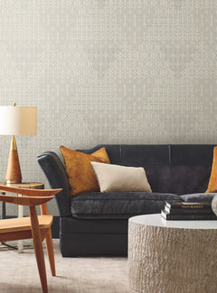 A modern living room featuring York Wallcoverings’ Modern Chandelier Wallpaper in soft gray tones, complemented by a dark gray sofa, gold and beige pillows, a round textured coffee table, a wooden end table, and a lamp with a gold base.
