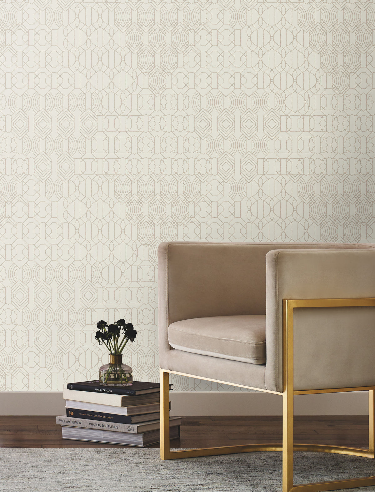 A sleek beige armchair with a gold frame sits beside a stack of books and a small vase on a rug, set against a wall adorned with the Modern Chandelier Wallpaper by York Wallcoverings.